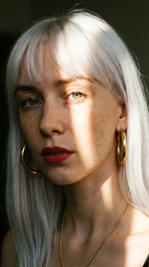 Intimate close-up portrait of young woman with white hair and freckles, pale green eyes, dramatic diagonal sunlight creating high contrast, gold hoop earrings, deep red lips