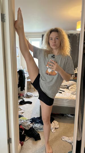 Photorealistic mirror selfie of Eastern European woman doing vertical standing split in messy bedroom, wearing graphic t-shirt and athletic shorts, natural lighting with iPhone