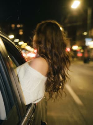 Vintage film photograph with dark grainy Kodak Portra look, woman leaning out of moving car window at night with heavy motion blur and wind-blown hair, dreamy melancholic atmosphere