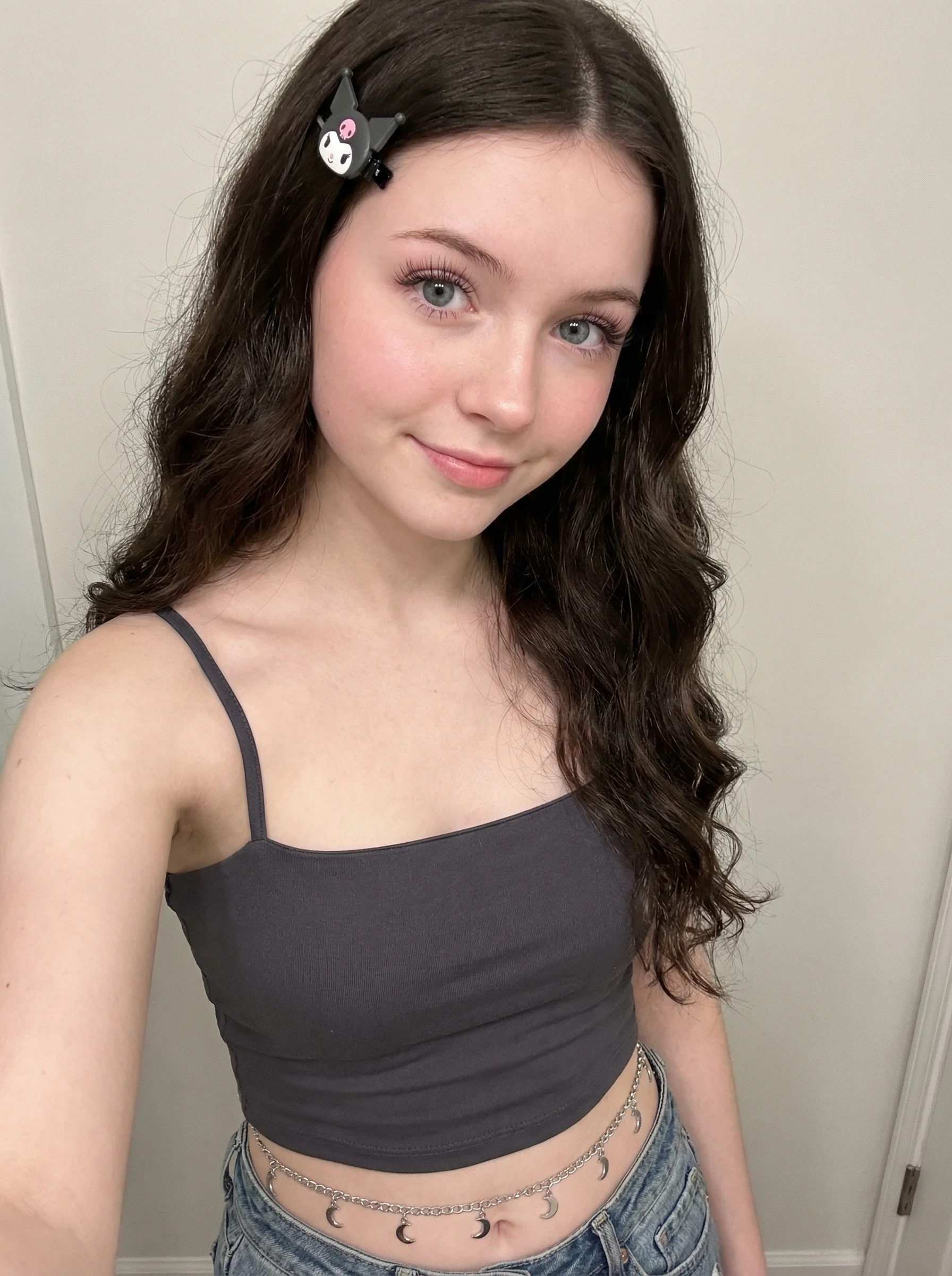 Teenage girl with dark brown wavy hair and Kuromi hair clip, wearing dark grey crop top with silver moon charm belt, soft pink makeup and blue-grey eyes