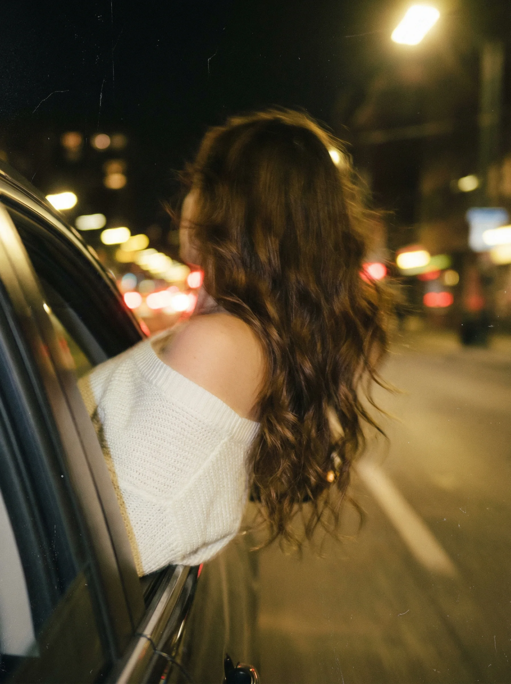 Vintage film photograph with dark grainy Kodak Portra look, woman leaning out of moving car window at night with heavy motion blur and wind-blown hair, dreamy melancholic atmosphere